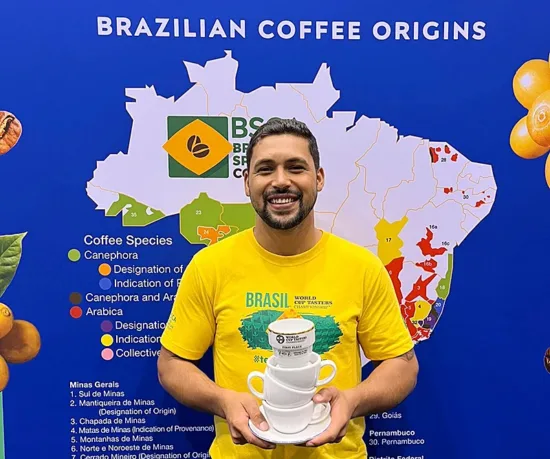 Dionatan in a yellow Brasil World Cup Tasters shirt, holding a trophy shaped like stacked coffee cups.