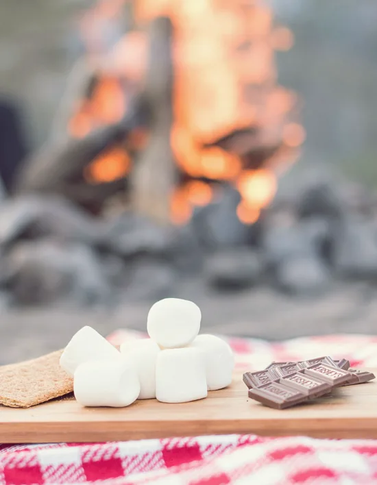 A wooden board in front of a campfire with graham crackers, chocolate, and marshmallows. 
