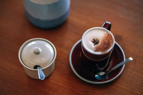 A mug and saucer on a table, inside which is a hot drink with ground cinnamon and a star anise pod for garnish. A sugar canister is next to it.