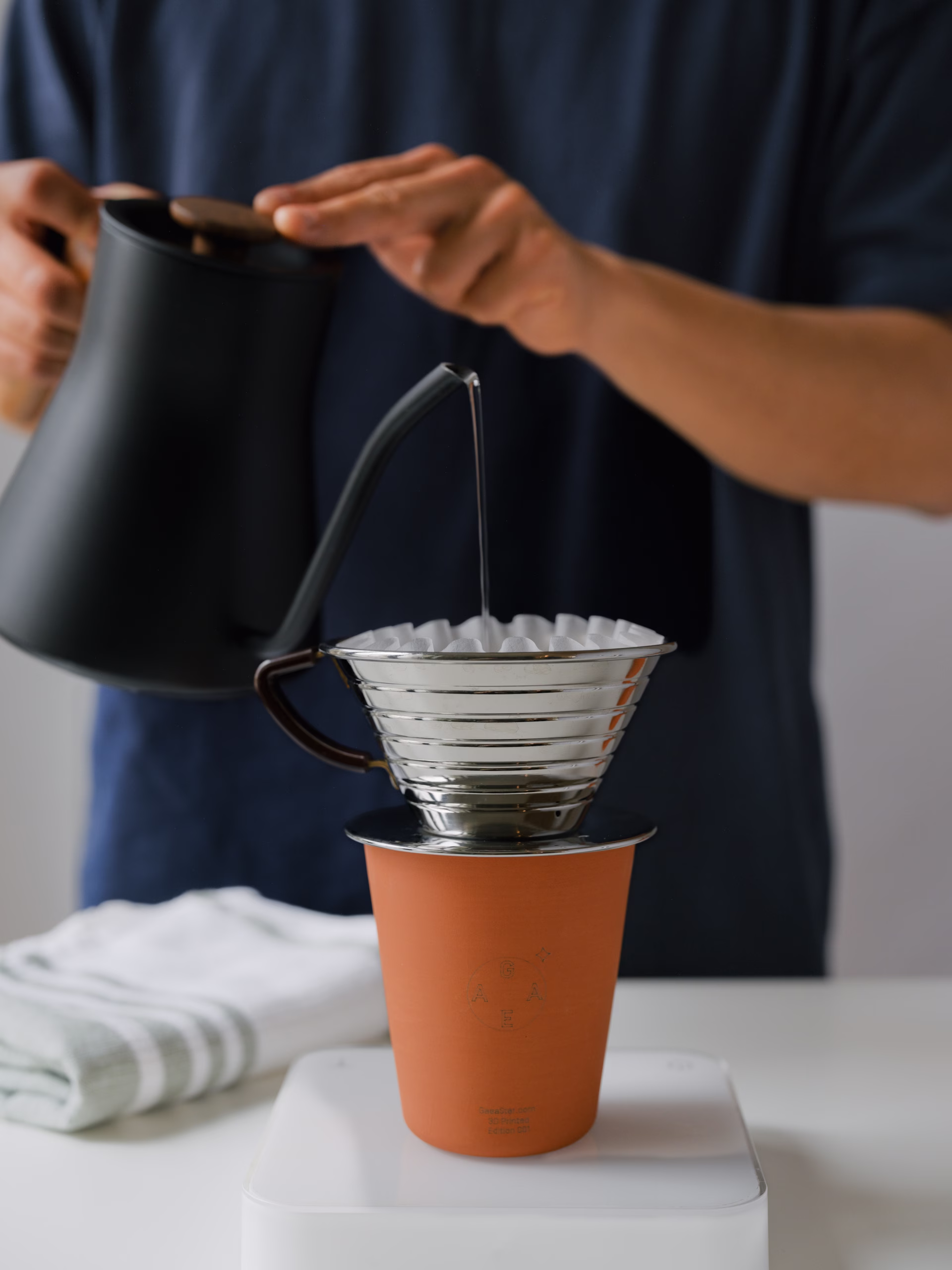 Un barista vierte agua caliente en un dispositivo de caída de café, que se encuentra encima de una taza de cerámica de color terracota por la marca GaeStar.