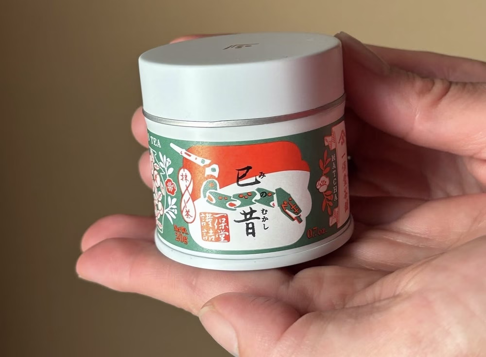 Un primer plano de una mano sosteniendo una lata de matcha decorada con imágenes florales.
