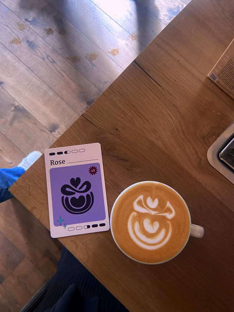 A card labeled “Rose” from the “Throw It Down” latte art game sits on a wooden table next to a mug filled with a coffee drink that is topped with rose-shaped latte art.